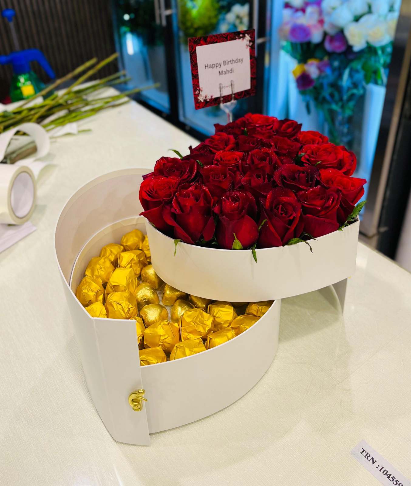 Heart-shaped box with red roses and gold chocolates on a table.